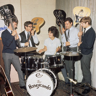 The Honeycombs Mark one line up, Honey Lantree seated at her custom built Carlton drum kit, surrounded by Martin Murray, Alan Ward, John Lantree, and Denis D'Ell, all holding guitars raised above her head. They are all smiling broadly.